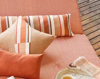 May include: A red and white herringbone patterned outdoor couch with a variety of pillows and a throw blanket. A white tray with a cup of tea and a sprig of dried flowers sits on the couch.