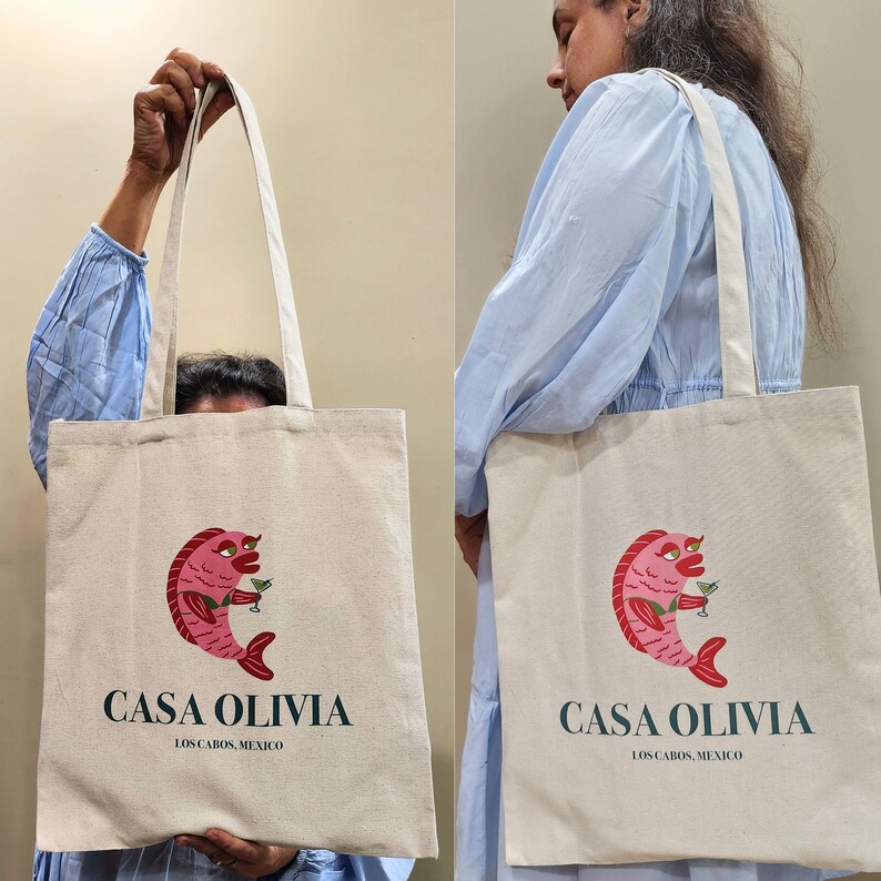 Puede incluir: Una bolsa de tela beige con una ilustraci&oacute;n de un pez rosa sosteniendo una bebida verde. La bolsa tiene el texto "Casa Olivia" y "Los Cabos, M&eacute;xico" impreso.