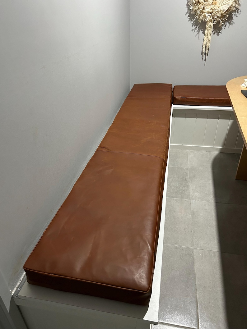 May include: Two brown leather bench cushions in a corner nook. The cushions are attached to a white bench.