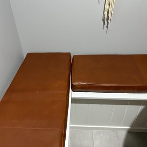 May include: Two brown leather cushions, one on top of the other, with a white wooden frame underneath. The cushions are in a corner of a room with a white wall and a white tiled floor.