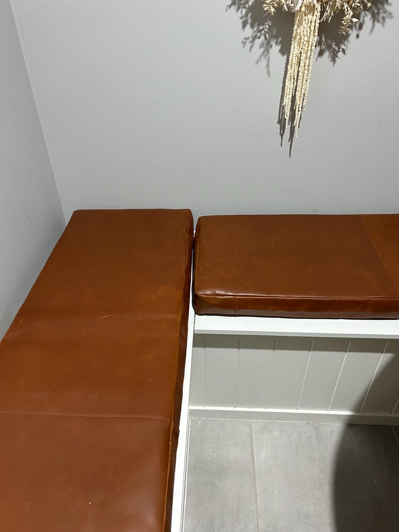 May include: Two brown leather cushions, one on top of the other, with a white wooden frame underneath. The cushions are in a corner of a room with a white wall and a white tiled floor.