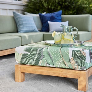 May include: A wooden ottoman with a green and white tropical leaf print fabric top. The ottoman is sitting on a gray concrete patio. A silver tray with two glass pitchers of lemonade and lemons sits on top of the ottoman.