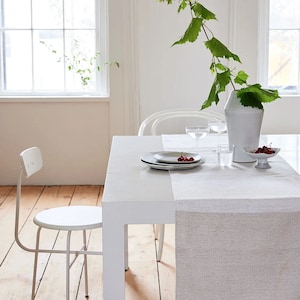 May include: A white dining table with a light gray runner is set with plates, cherries, and glassware. A white vase holds a green leafy branch. A white chair is positioned to the side. The room has a minimalist, modern aesthetic.