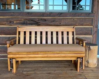 May include: A wooden glider bench with a brown cushion and armrests. The bench has a slatted back and is made of light-coloured wood. A wooden stump side table is next to the bench.