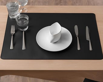 May include: A black leather placemat with a white ceramic cup and saucer, two silver forks, and two silver knives.