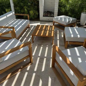 Puede incluir: Juego de muebles de patio exterior de madera clara con cojines blancos. El conjunto incluye un sofá, dos sillones, dos otomanas y una mesa de centro cuadrada. Los muebles están colocados sobre un suelo de piedra, con sombras.