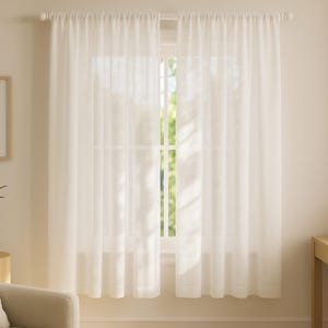 May include: Light cream-coloured curtains hang in front of a window, allowing natural light to filter through. The curtains are hung on a white rod. A framed abstract art piece and a small potted plant are visible.