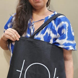 Puede incluir: Una mujer con un vestido estampado en azul y blanco sostiene una bolsa de mano negra con un c&iacute;rculo blanco y l&iacute;neas que forman la palabra "TOK" en ella.