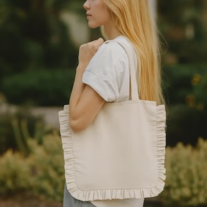 May include: Cream-colored tote bag with a ruffled edge. The bag has a rectangular shape with a simple design and long straps for carrying over the shoulder. The bag is made of a soft, neutral-toned fabric.