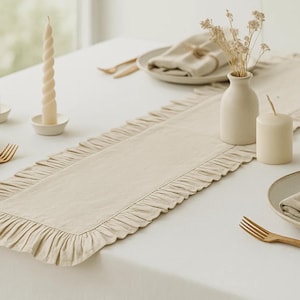 May include: A beige linen table runner with a ruffled edge, set on a white tablecloth. The table is decorated with candles, plates, napkins, and wooden forks, creating a warm, inviting dining scene.
