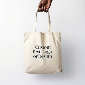Puede incluir: Una bolsa de lona blanca con texto negro que dice "Custom Text, Logo, or Design".