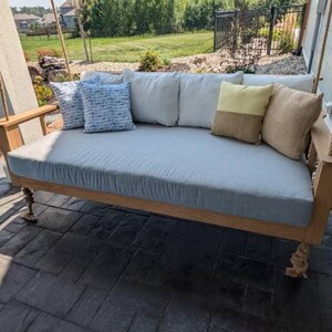 May include: A wooden swing bed with a light blue cushion and a variety of patterned pillows. The swing bed is hanging from a rope and is in a backyard setting.