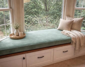 Puede incluir: Un asiento de ventana incorporado con un cojín verde azulado, dos almohadas beige y una manta con flecos. Una pequeña bandeja con un jarrón de flores y una vela se encuentra en el asiento. La ventana da a un bosque verde.