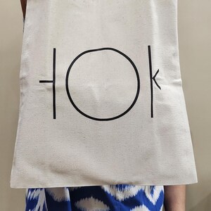 Puede incluir: Una bolsa de lona blanca con asas negras y la palabra "TOK" impresa en negro en la parte delantera. La "O" de la palabra es un c&iacute;rculo.