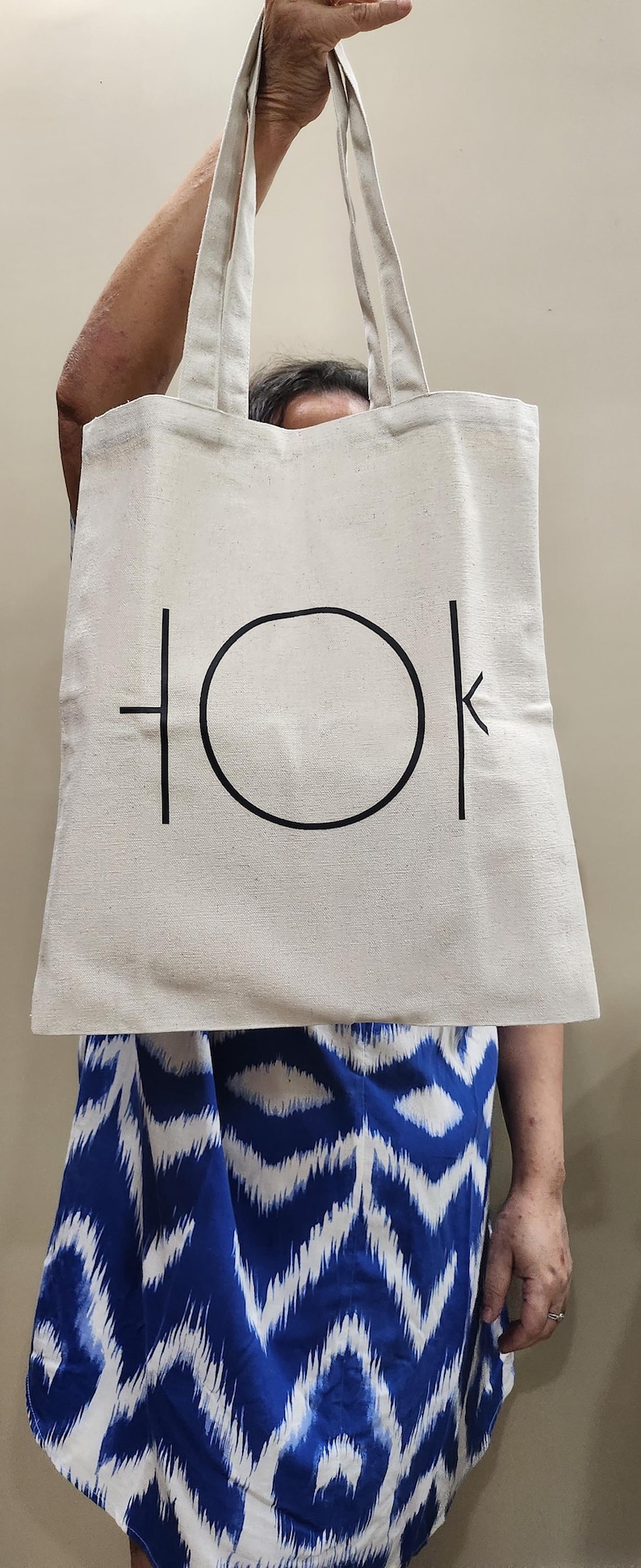 Puede incluir: Una bolsa de lona blanca con asas negras y la palabra "TOK" impresa en negro en la parte delantera. La "O" de la palabra es un c&iacute;rculo.