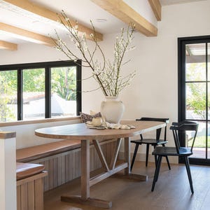 May include: A light wood dining table with a vase of white flowers in a bright room. Two black chairs are positioned near the table. A built-in bench with a leather cushion is on the left.