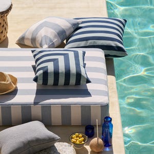 Puede incluir: Una escena junto a la piscina con cojines a rayas y una tumbona. Los cojines tienen rayas azules y blancas y diseños geométricos. También hay una mesa auxiliar tejida y objetos decorativos.