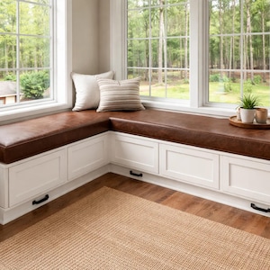May include: A built-in window seat with a brown leather cushion and white storage cabinets. Two pillows rest on the seat, and a small tray with plants sits on the right. The window overlooks a lush green forest.