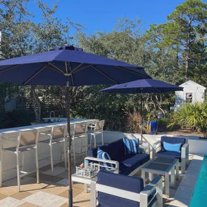 May include: An outdoor patio scene featuring navy blue parasols, a white bar with beige bar stools, and a seating area with a navy blue sofa, chairs, and small tables. The background includes trees and a clear blue sky.