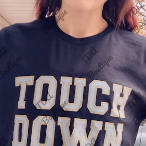 May include: A black t-shirt with the words "TOUCH DOWN" in white and gold glitter lettering. The shirt is worn by a person with brown hair and glasses.