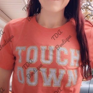 May include: A coral colored t-shirt with the words "TOUCHDOWN" in silver glitter letters. The shirt has the text "TDA Boutique" printed on the front and back.