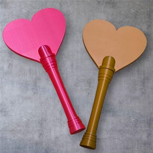 May include: Two heart-shaped hand fans. One is bright pink, the other is tan. Both have long, cylindrical handles. The pink fan's handle is red, and the tan fan's handle is olive green. The fans are made of a textured material.