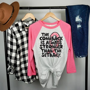 May include: A pink and grey baseball tee shirt with a black graphic that reads "The Comeback is Always Stronger Than The Setback" with a pink ribbon graphic.
