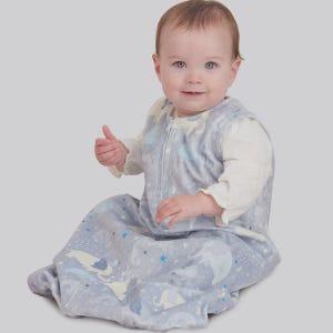 May include: A baby in a light grey sleep sack featuring a unicorn and star design. The sleep sack has a zip closure and is worn over a white long-sleeved top. The baby is sitting and smiling.