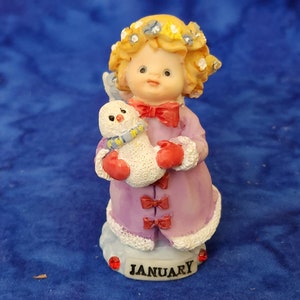 May include: A purple-clad angel figurine with blonde hair and a flower crown holds a small snowman. The base of the figurine is white with the word "JANUARY" in black lettering.