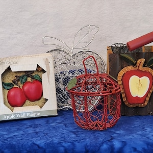 May include: Three apple-themed wall decorations. A red wire apple basket, a silver wire apple, and a wooden plaque with the words "Gift Gallery" and "Apple Wall Plaque" with a painted image of two red apples.