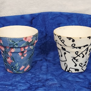 May include: Two small white ceramic flower pots. One pot is decorated with a blue and pink floral pattern. The other pot is decorated with a black and white musical note pattern.