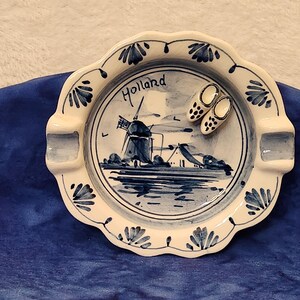 May include: A white ceramic ashtray with a blue and white Delft design featuring a windmill and the word "Holland". Two small white and blue clogs are on the rim of the ashtray.