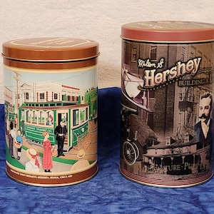 May include: Two vintage Hershey's chocolate tins. One cylindrical tin features a green trolley scene with people and the text "Chocolate Town Trolleys, Hershey, Penna, Circa 1920." The other tin has a sepia-toned image of the Hershey Building.