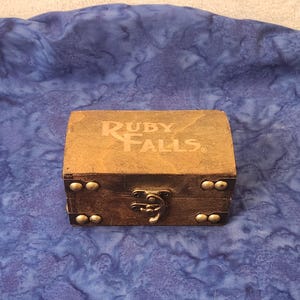 May include: A small, rectangular wooden box with the words "RUBY FALLS" printed on the top. The box is brown with brass-colored studs and a clasp. The background is a blue fabric.
