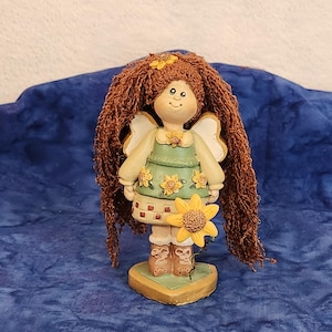 May include: A figurine of a girl with long brown hair, wearing a green dress with yellow flowers and a sunflower in her hand. She is standing on a heart-shaped base.
