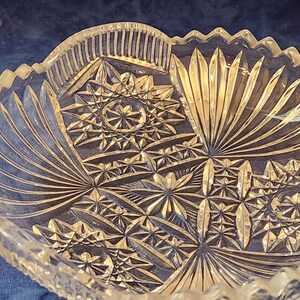 May include: A clear glass bowl with detailed starburst and fan patterns. The bowl has a scalloped edge and a textured surface. The design features geometric shapes and radial lines, creating a decorative appearance. A vintage piece, suitable for serving.
