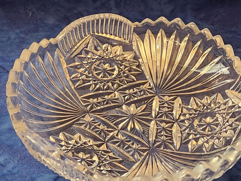 May include: A clear glass bowl with detailed starburst and fan patterns. The bowl has a scalloped edge and a textured surface. The design features geometric shapes and radial lines, creating a decorative appearance. A vintage piece, suitable for serving.