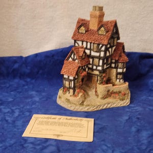 May include: A miniature decorative house with a red tile roof, brown chimney, and white and brown walls. The house sits on a beige base with small red flowers. A certificate of authenticity is in front of the house.