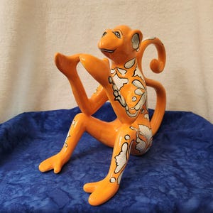 May include: Orange ceramic monkey figurine with white and black floral patterns. The monkey is in a seated position with its arms and legs extended. The figurine is on a blue fabric surface.