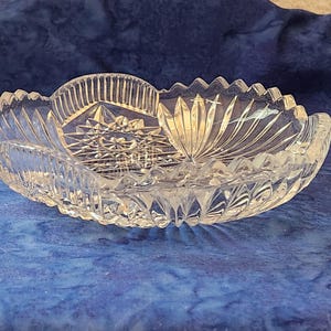 May include: A clear, oval crystal bowl featuring starburst and fan patterns. The bowl has a scalloped edge and is designed with detailed cuts, reflecting light. This decorative piece is suitable for serving or display.