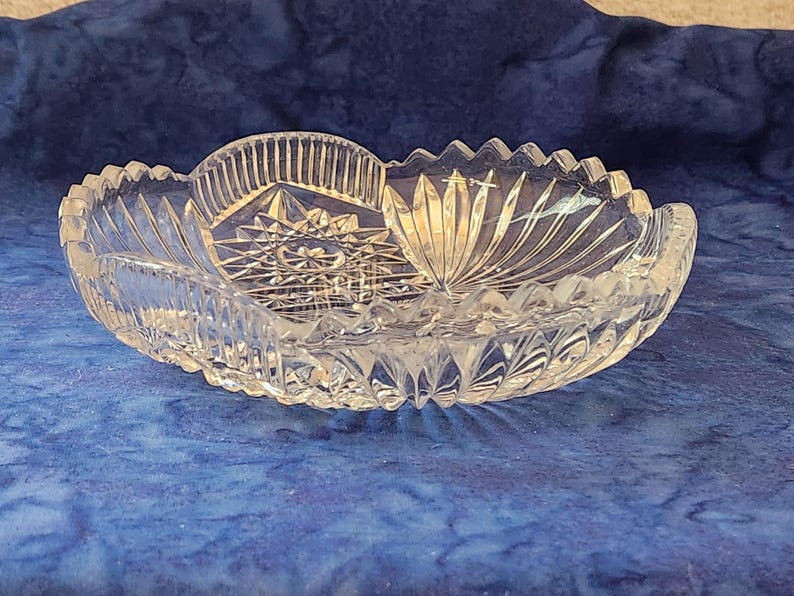 May include: A clear, oval crystal bowl featuring starburst and fan patterns. The bowl has a scalloped edge and is designed with detailed cuts, reflecting light. This decorative piece is suitable for serving or display.