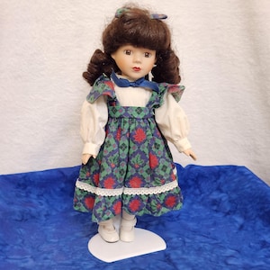 May include: A porcelain doll with brown curly hair and a blue hair bow. The doll wears a white long-sleeved blouse with a blue ribbon tie, a green, red, and blue plaid jumper dress with white lace trim at the hem, and white shoes.