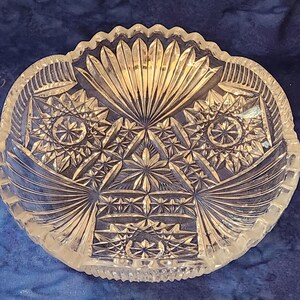 May include: Clear glass serving bowl with intricate starburst and geometric patterns. The bowl has a scalloped edge and a symmetrical design, ideal for displaying fruit or serving snacks. The bowl's design is reminiscent of vintage glassware.