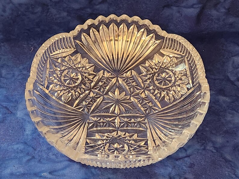 May include: Clear glass serving bowl with intricate starburst and geometric patterns. The bowl has a scalloped edge and a symmetrical design, ideal for displaying fruit or serving snacks. The bowl's design is reminiscent of vintage glassware.