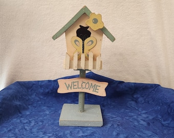 May include: A wooden decorative birdhouse with a butterfly design and a welcome sign. The birdhouse has a light pink body, a green roof, and a yellow flower accent. The sign reads "WELCOME".