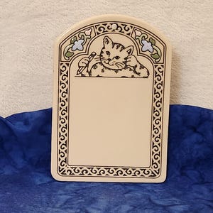 May include: Cream-colored ceramic plaque with a decorative border and a cat illustration. The cat is holding a pencil. The top border has floral and leaf accents in blue, green, and black. The plaque is on a blue surface.
