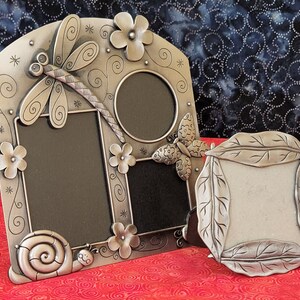 May include: A silver-toned picture frame with a dragonfly, flowers, and a snail design. The frame has three openings for pictures. There is a smaller silver-toned picture frame with a leaf design.