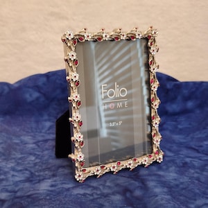 May include: A rectangular silver-toned picture frame with a decorative border featuring small white flowers and red crystal accents. The glass insert displays the text Folio HOME and 3.5 x 5 inches. The frame has a black easel back support.
