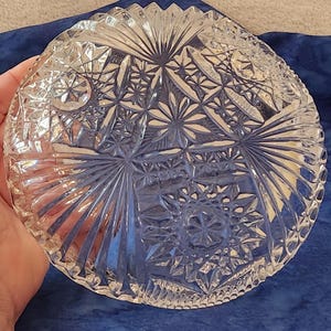 May include: A clear, round glass bowl with intricate starburst and geometric patterns. The bowl has a scalloped edge and is held in a hand, showcasing the detailed cut glass design. The bowl is likely for serving or display.