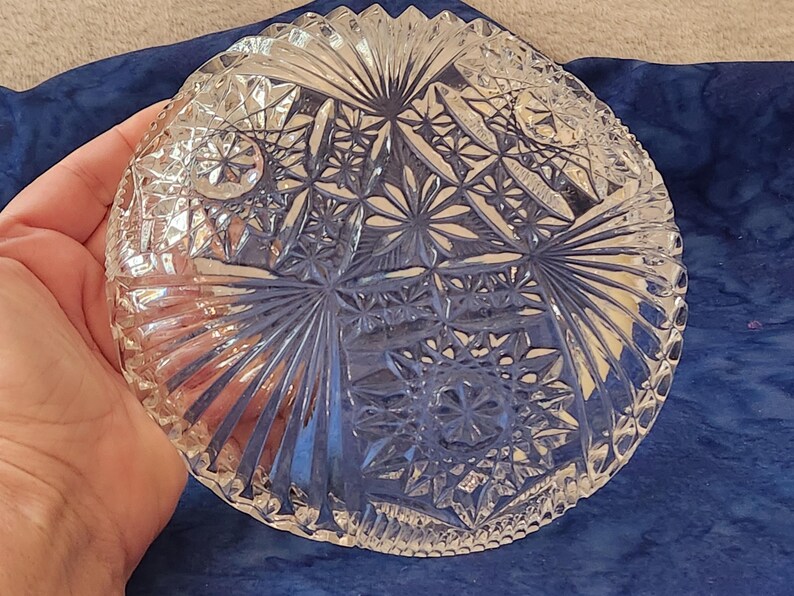 May include: A clear, round glass bowl with intricate starburst and geometric patterns. The bowl has a scalloped edge and is held in a hand, showcasing the detailed cut glass design. The bowl is likely for serving or display.